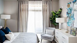 A modern bedroom with a bed, a chair, a dresser, and a large window with curtains. - Kings Point Lux Suites (Great Neck)