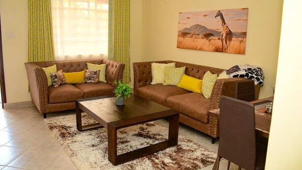 Un salon avec deux canapés marron, une table basse, une table à manger avec des chaises et un tableau représentant une girafe accroché au mur.