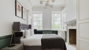 A bedroom with a large bed, a bedside table with a lamp, a fireplace, and framed artwork on the wall.