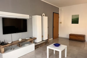 A modern living room with a flat-screen TV, wooden and white furniture, a small white table, and a door. - Amarea holiday home in Fuscaldo (Marina di Fuscaldo)