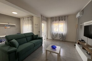 Un salon avec un canapé vert, une petite table blanche sur laquelle se trouve un bol bleu, un téléviseur à écran plat et une étagère en bois.