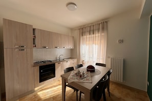 A compact kitchen with wooden cabinets, a gas stove, and a dining area with a table set for two. - Amarea holiday home in Fuscaldo (Marina di Fuscaldo)