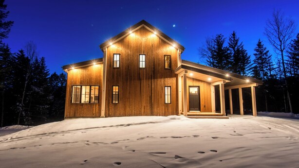 A two-story wooden house with a covered porch and multiple windows, surrounded by snow and trees. - Golden Hour: 3-bedroom luxury home in the Canadian Wilderness (Golden)