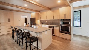 A modern kitchen with wooden cabinets, a central island with bar stools, stainless steel appliances, and a window with a view. - Golden Hour: 3-bedroom luxury home in the Canadian Wilderness (Golden)