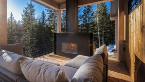 A cozy outdoor seating area with a fireplace, wooden deck, and a view of a forest. - Golden Hour: 3-bedroom luxury home in the Canadian Wilderness (Golden)