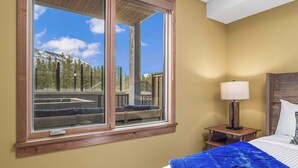 Quarto com uma janela ampla, cama com manta azul, mesa de cabeceira de madeira com abajur, e vista para um deck e montanhas.