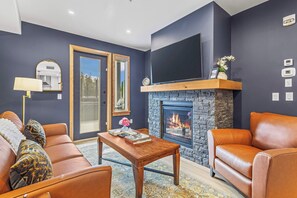 A modern living room with a fireplace, leather sofas, a coffee table, and a flat-screen TV mounted above the fireplace. - Spring Creek Vacations 1 BR Condo 106TM (Canmore)