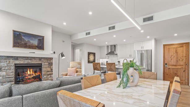 A modern living room with a fireplace, grey sofas, a marble dining table, and a kitchen in the background. - Spring Creek Vacations 2 BR Premier Condo 109TM (Canmore)