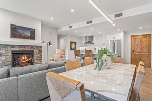 Una sala de estar moderna con chimenea, sofás grises, una mesa de comedor de mármol y una cocina al fondo.