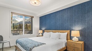 A bedroom with a large bed, two bedside tables with lamps, a window with a view of mountains, and a blue textured wall. - Spring Creek Vacations 2 BR Premier Condo 109TM (Canmore)