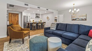 Una sala moderna con sofá, reposapiés y una silla. Hay un comedor con sillas y una mesa, además de una cocina con refrigerador y estufa.