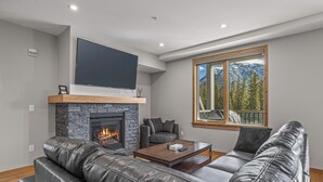 A modern living room with a fireplace, leather sofa, and a large window with a mountain view. - Spring Creek Vacations 3 BR Premier Condo 210TM (Canmore)