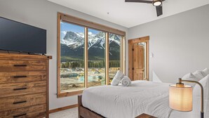 A bedroom with a large window offering a mountain view, a wooden dresser, a bed with white linens, and a bedside lamp. - Spring Creek Vacations 3 BR Premier Condo 210TM (Canmore)