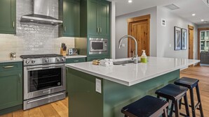 A modern kitchen with green cabinets, stainless steel appliances, a large island with stools, and a wooden door. - Spring Creek Vacations 3 BR Premier Condo 210TM (Canmore)