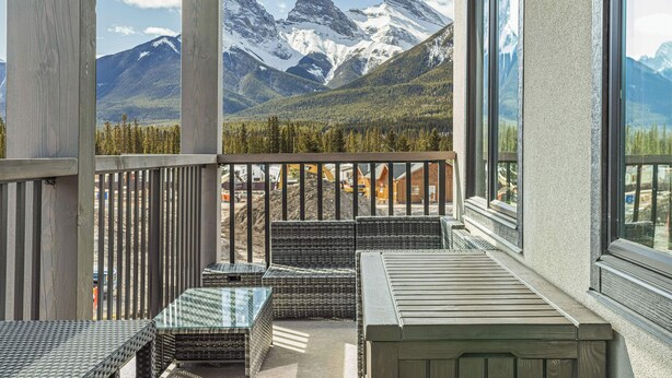 A balcony with wicker furniture and a glass table, overlooking a mountainous landscape with snow-capped peaks. - Spring Creek Vacations 3 BR Premier Condo 210TM (Canmore)