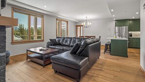 A modern living room with a leather sofa, wooden coffee table, and a view of the outdoors through large windows. - Spring Creek Vacations 3 BR Premier Condo 210TM (Canmore)