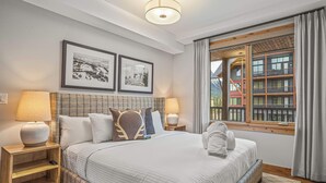 A neatly arranged bedroom with a large bed, two bedside tables with lamps, framed pictures on the wall, and a view of a building through a window. - Spring Creek Vacations 3 BR Premier Condo 213TM (Canmore)