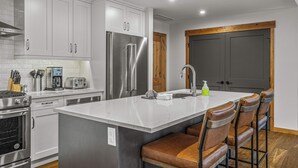A modern kitchen with white cabinetry, stainless steel appliances, and a marble island with bar stools. - Spring Creek Vacations 3 BR Premier Condo 213TM (Canmore)