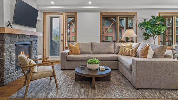 A modern living room with a stone fireplace, a sectional sofa, a wooden chair, a coffee table with a plant, and a TV mounted on the wall. - Spring Creek Vacations 3 BR Premier Condo 213TM (Canmore)