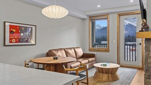 A modern living room with a leather sofa, wooden dining table, and chairs, a round coffee table, a pendant light, and a framed artwork on the wall. - Spring Creek Vacations 1 BR Condo 211TM (Canmore)