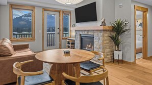 A cozy living room with a fireplace, wooden furniture, and large windows offering mountain views. - Spring Creek Vacations 1 BR Condo 211TM (Canmore)