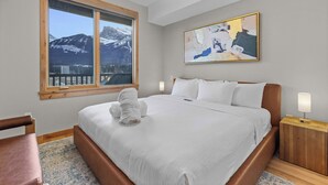 A hotel room with a large bed, a wooden headboard, a nightstand with a lamp, a painting on the wall, and a window with a mountain view. - Spring Creek Vacations 1 BR Condo 211TM (Canmore)