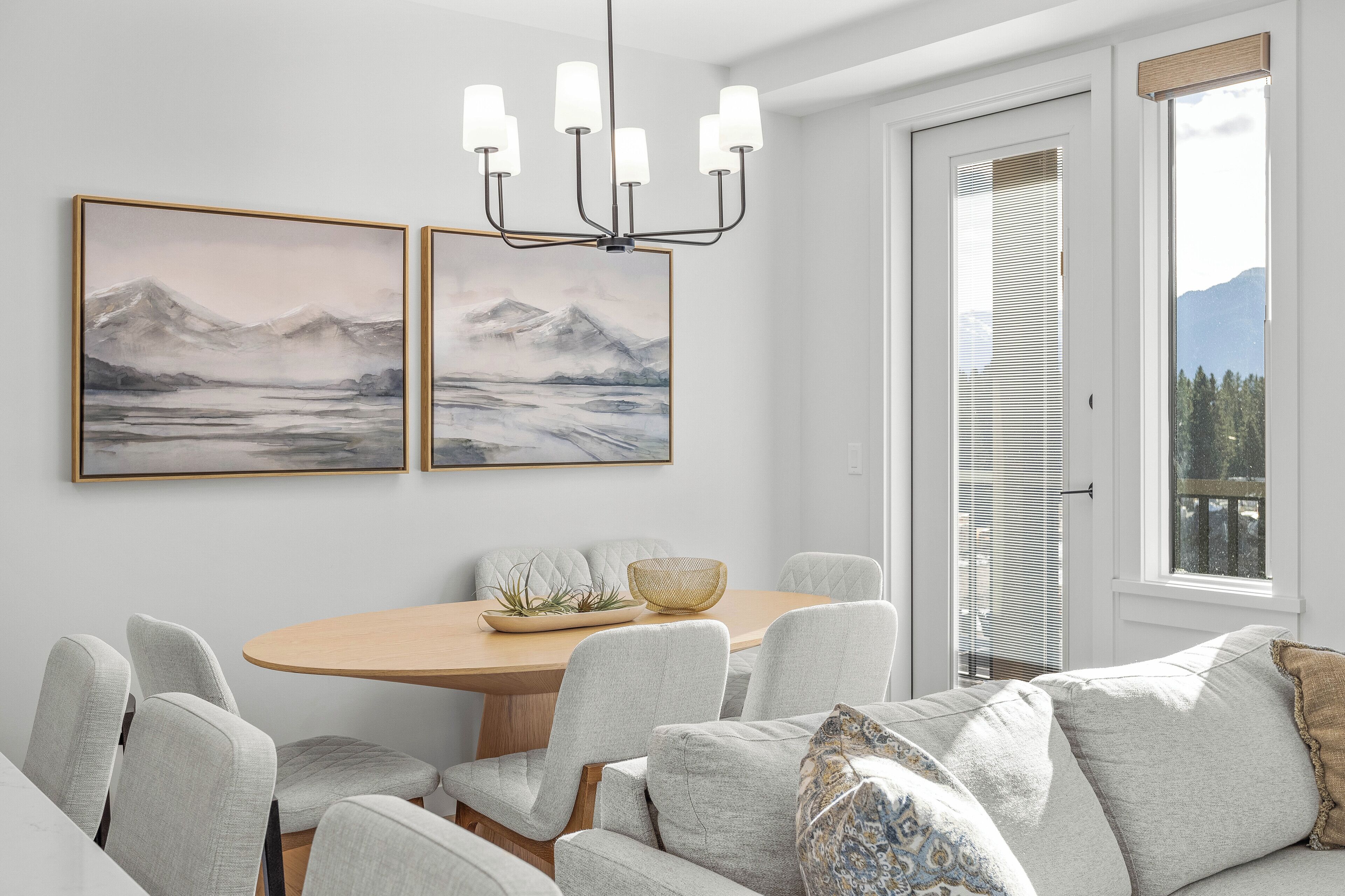 A dining area with a round table, grey upholstered chairs, and a chandelier. There are two framed paintings on the wall and a window with blinds.