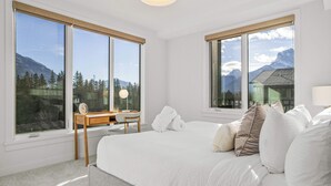 A bedroom with a large bed, two windows with mountain views, a bedside table with a lamp, and a view of a mountain cabin. - Spring Creek Vacations 2 BR Premier Condo 304TM (Canmore)