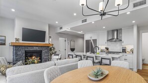 A modern living room with a fireplace, grey sofas, a dining table, and a kitchen area with white cabinets and stainless steel appliances. - Spring Creek Vacations 2 BR Premier Condo 304TM (Canmore)
