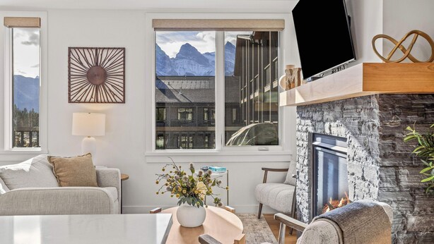 A modern living room with a fireplace, a large window with a mountain view, and a flat-screen TV mounted above the fireplace. - Spring Creek Vacations 2 BR Premier Condo 304TM (Canmore)