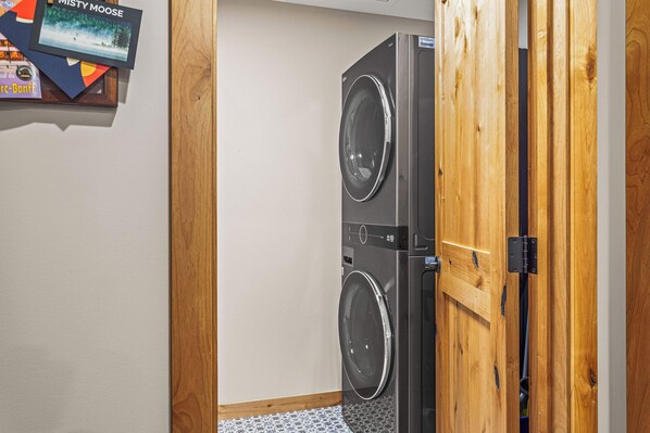 Une buanderie équipée de deux machines à laver et deux sèche-linge superposés, de portes en bois et d’une petite étagère où sont rangés divers objets.