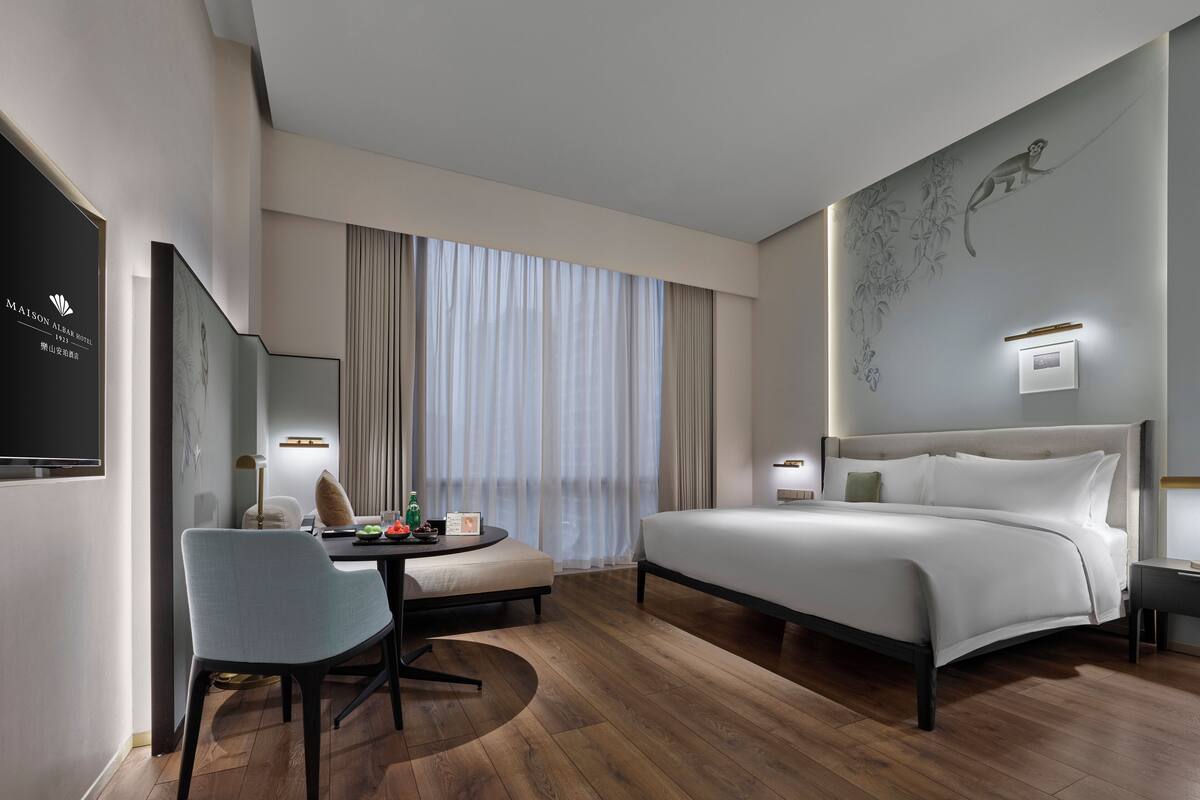 A modern hotel room with a bed, a desk, a chair, a dining table, and a large window with curtains.