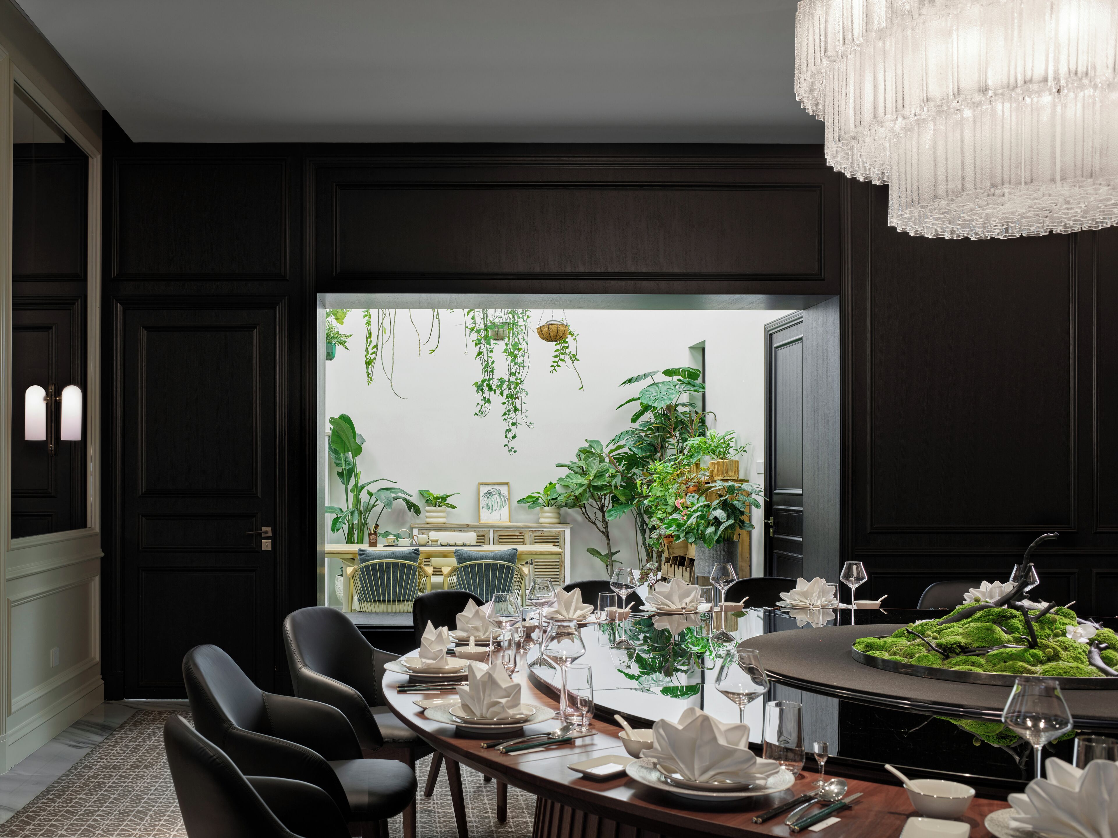 A dining area with a dark wood panel wall, a large window with plants, a chandelier, and a table set for a meal.