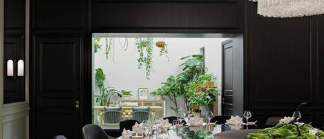 A dining area with a dark wood panel wall, a large window with plants, a chandelier, and a table set for a meal.