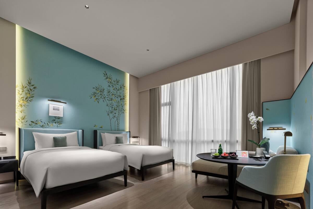 A modern hotel room with two beds, a dining area, and a large window with curtains.