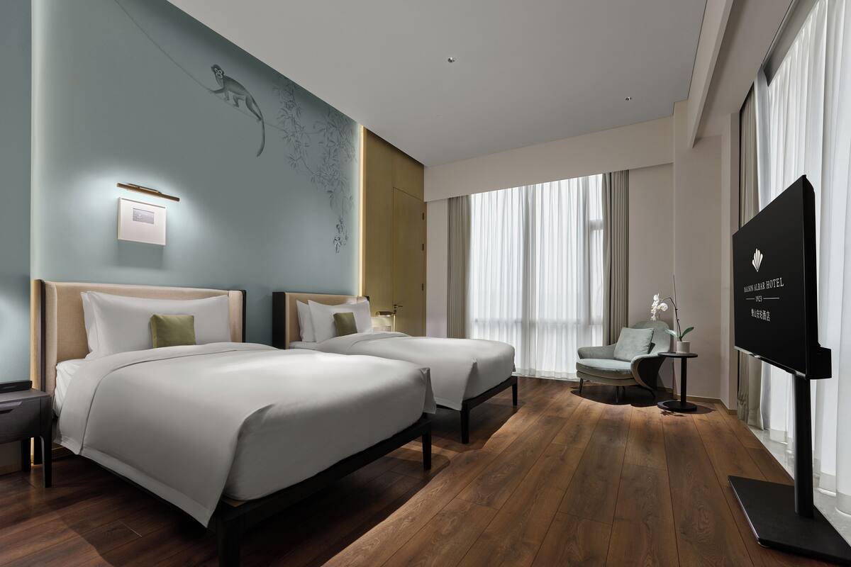 A hotel room with two beds, a desk, a chair, a TV, and a large window with curtains.