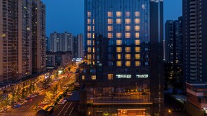 Un hotel moderno con fachada de vidrio, rodeado de rascacielos y una calle concurrida al anochecer.