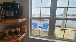 Et køkken med kaffemaskine, krukker og et vindue med udsigt over en bro og nogle stole.