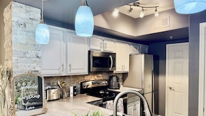 A modern kitchen with white cabinets, a stone accent wall, a built-in microwave, a stainless steel refrigerator, and pendant lighting. - Modern North Scottsdale Condo – Resort Style Amenities, 1st Floor Corner Unit (Scottsdale)