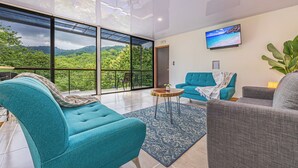 A modern living room with a large glass door leading to a balcony, a flat-screen TV mounted on the wall, and a blue and gray sofa set. - 3BR - Mountain View - Amazing Pool - 5 min to the Beach - Guanacaste (Puerto Carrillo)