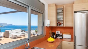 Una cocina moderna con un lavamanos de acero inoxidable, un refrigerador Siemens y una cafetera. Se aprecia vista al mar a través de amplias ventanas.