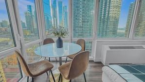 A modern room with a round glass table, four chairs, a vase with flowers, and a view of a cityscape through large windows.