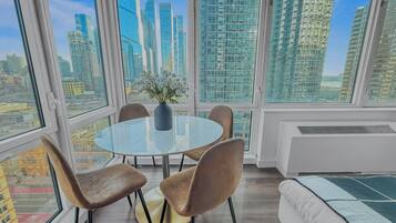 Een moderne kamer met een ronde glazen tafel, vier stoelen, een vaas met bloemen en een stadsgezicht door grote ramen.