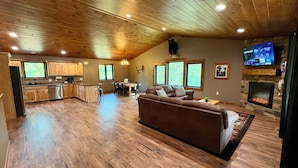 A spacious living area with a wooden ceiling, a fireplace, a television, and a kitchen in the background. - Bear Paw Lodge • Luxury Cabin • Game Room • Fire Pit (Wisconsin Dells)