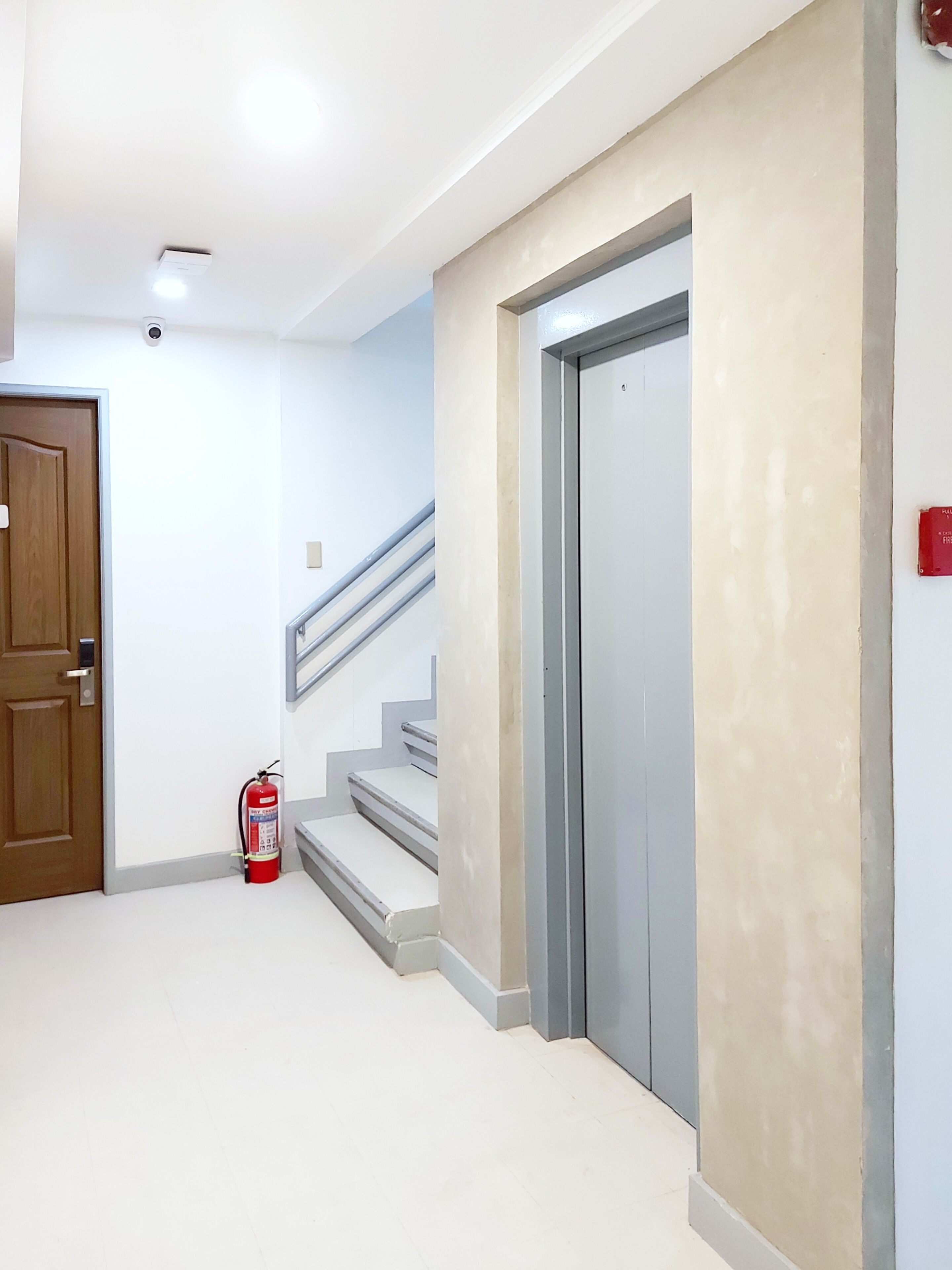 Um corredor com uma porta de madeira, um extintor de incêndio e uma escada que dá acesso a um elevador.