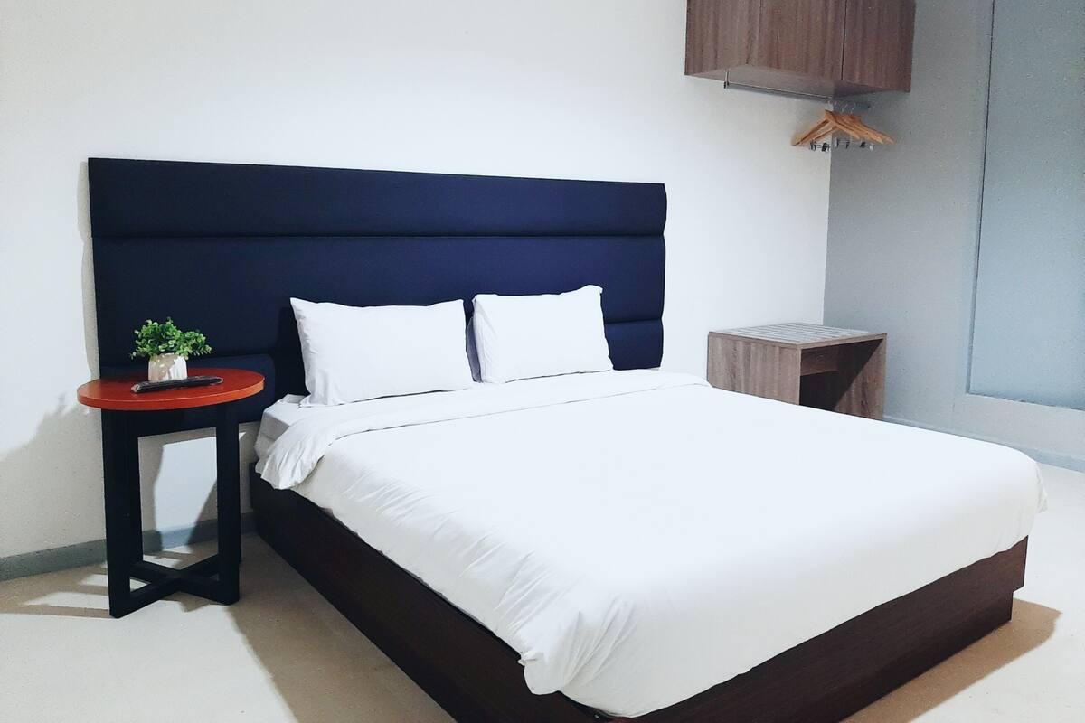 A neatly made bed with white linens, a dark blue upholstered headboard, and a small round table with a potted plant beside it.
