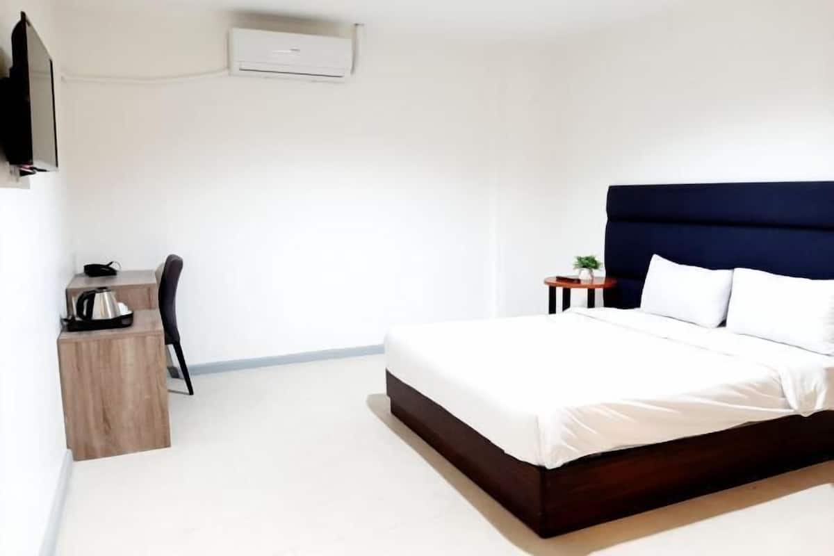 A bedroom with a bed, a desk, a chair, a small table with a plant, and an air conditioning unit on the wall.