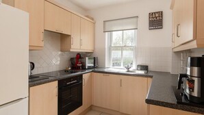 Een moderne keuken met lichte houten kastjes, een zwarte oven, een magnetron, een koffiezetapparaat en een raam met jaloezieën.