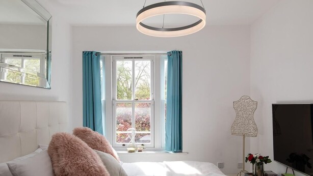 Een moderne slaapkamer met een bed, een groot raam met gordijnen, een nachtkastje met een lamp en een televisie.