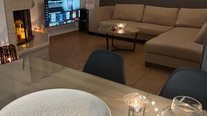 Ein modernes Wohnzimmer mit einem beigefarbenen Sofa, einem Glastisch und einem Esstisch mit dekorativer Platte. Ein Flachbildfernseher ist an der Wand montiert, und es gibt einen Kamin mit einer brennenden Kerze auf dem Tisch.
